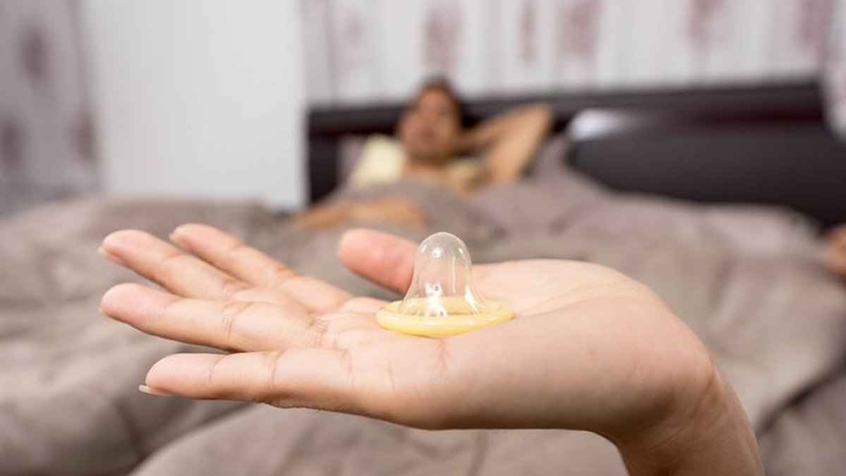En hand håller upp en kondom. I bakgrunden ligger en man i sängen.