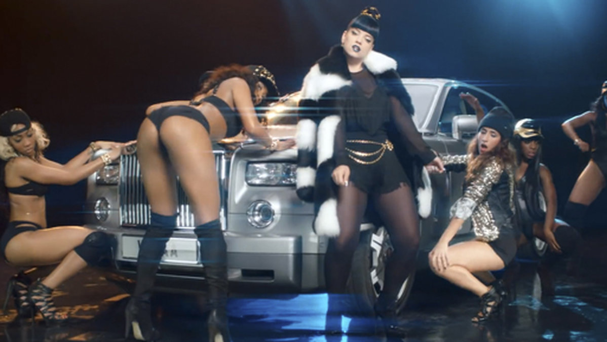 En påklädd Lily Allen flankerad av dansöser i videon.