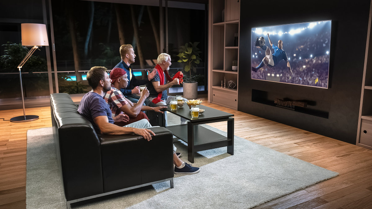 Flera män sitter i en soffa och tittar på fotboll på en TV